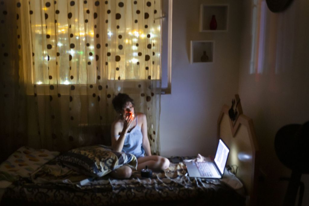 Une jeune femme fume sur son lit en regardant son ordinateur le soir