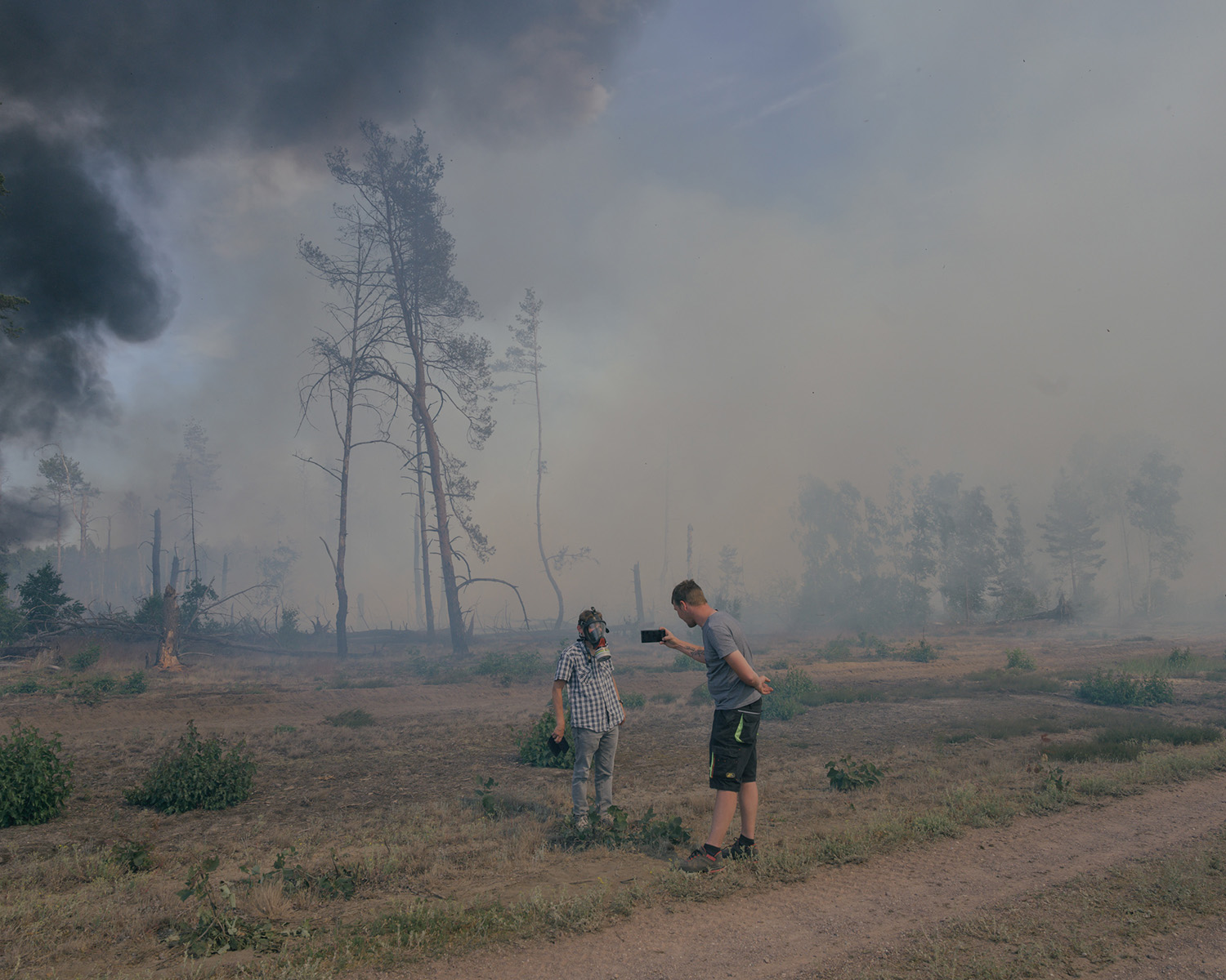 Sur fond de feu de forêt, une homme prend en photo un autre homme portant un masque à gaz